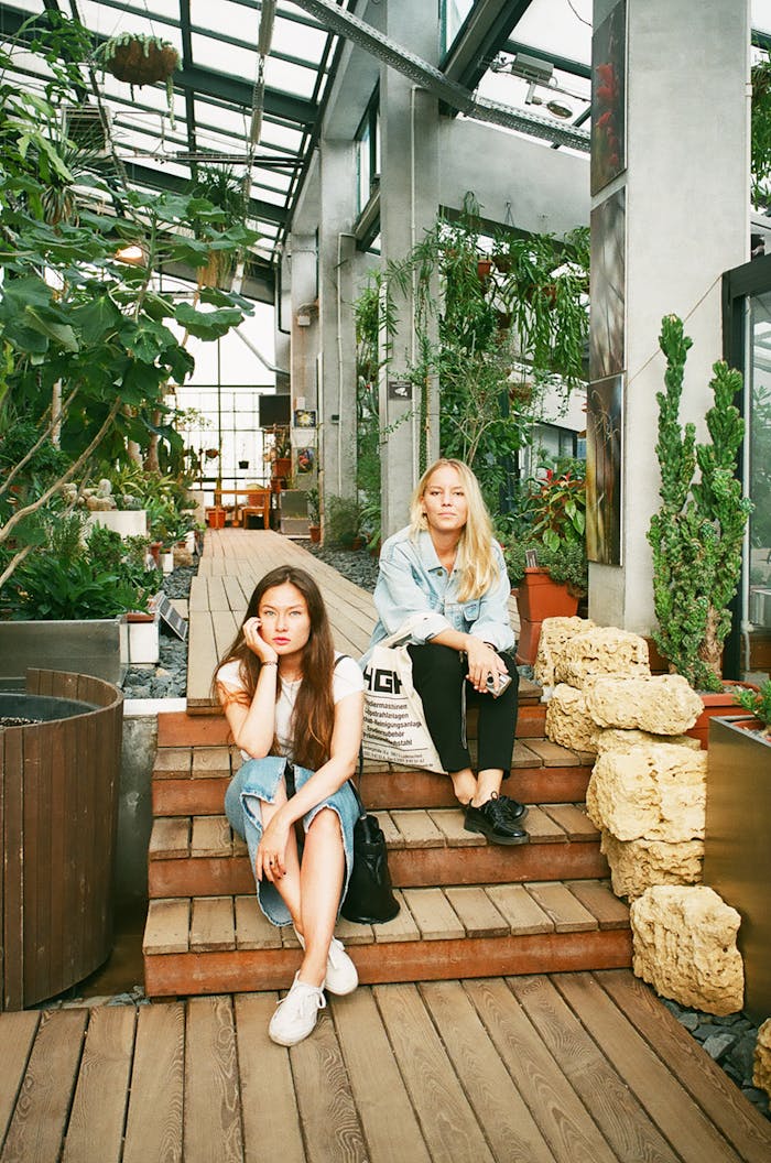gallery-2 Two women sitting on wooden steps in a modern greenhouse, showcasing casual fashion.